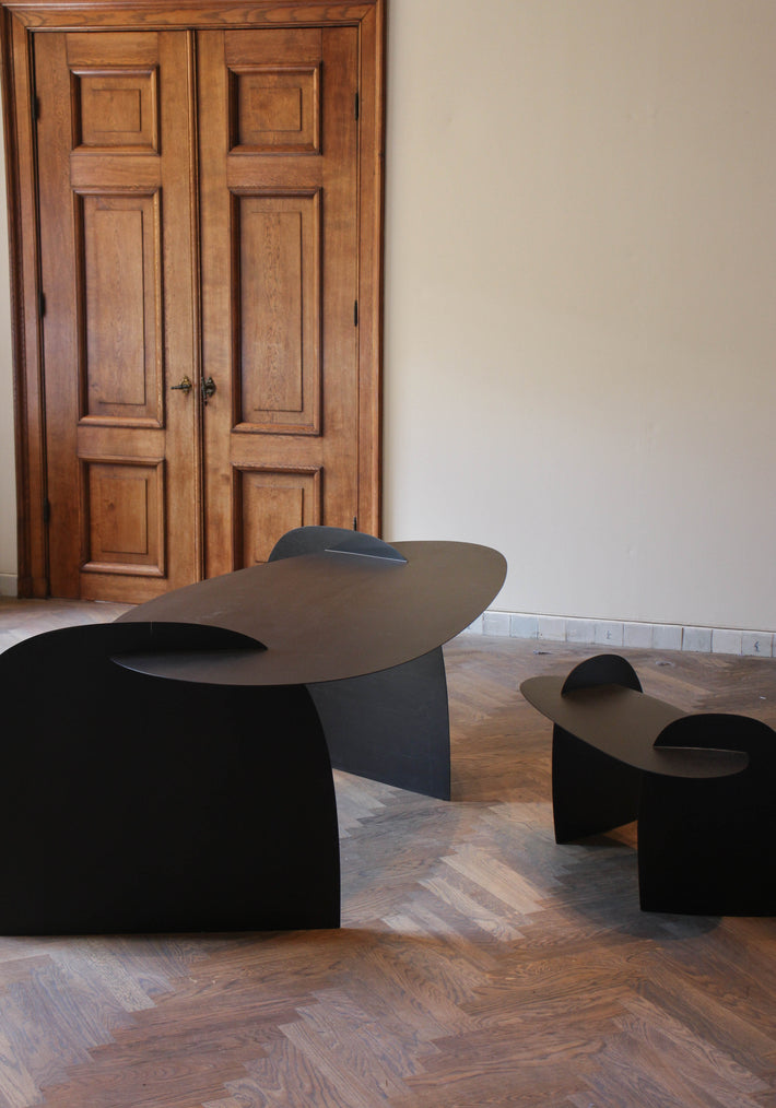 Sculpted Table | 9845: Matte-black oak table with curved, interlocking panels in a minimalist design, set in a room with herringbone wood flooring and tall doors.