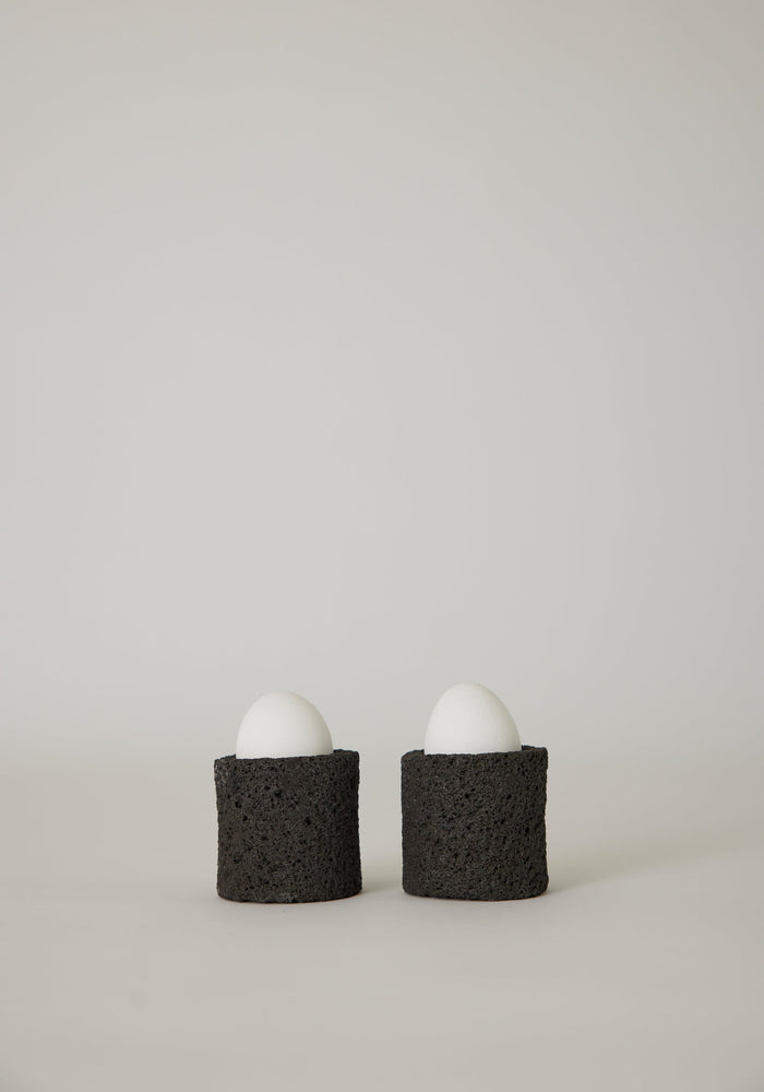Egg Cups: Two white eggs in handcrafted black holders made from lava stone, showcasing a unique porous texture, designed by artist Louis Crombag.
