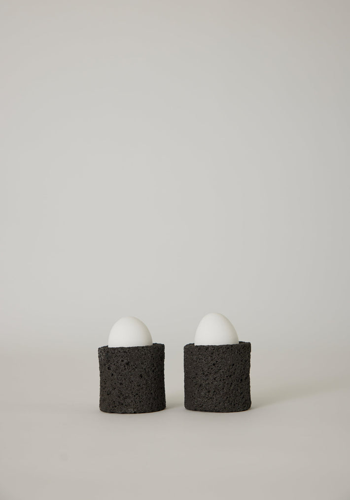 Egg Cups: Two white eggs in handcrafted black holders made from lava stone, showcasing a unique porous texture, designed by artist Louis Crombag.