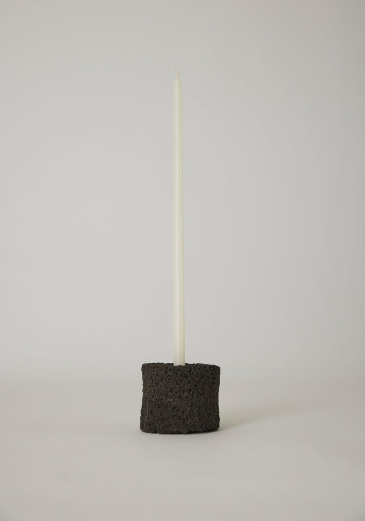 Round Candle Holder: Slim white taper candle in a handcrafted black lava stone holder, showcasing minimalist design and artisanal craftsmanship by Dutch artist Louis Crombag.