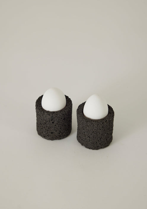 Egg Cups: Two handcrafted lava stone cups holding eggs, set on a linen napkin with a decorative teaspoon, showcasing unique craftsmanship by Dutch artist Louis Crombag.