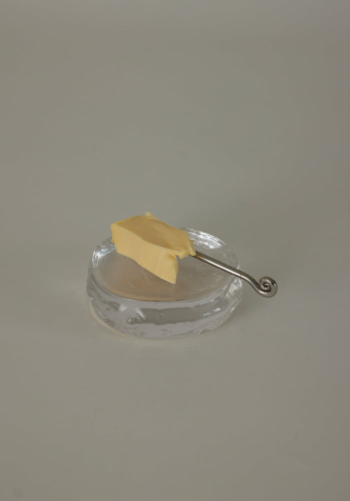 Butter plate | Glass by Szkło Studio: An irregular clear glass dish with a silver spreader knife and butter pat, showcasing unique, hand-blown craftsmanship.