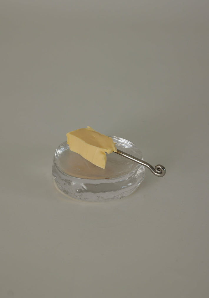 Butter plate | Glass by Szkło Studio: An irregular clear glass dish with a silver spreader knife and butter pat, showcasing unique, hand-blown craftsmanship.