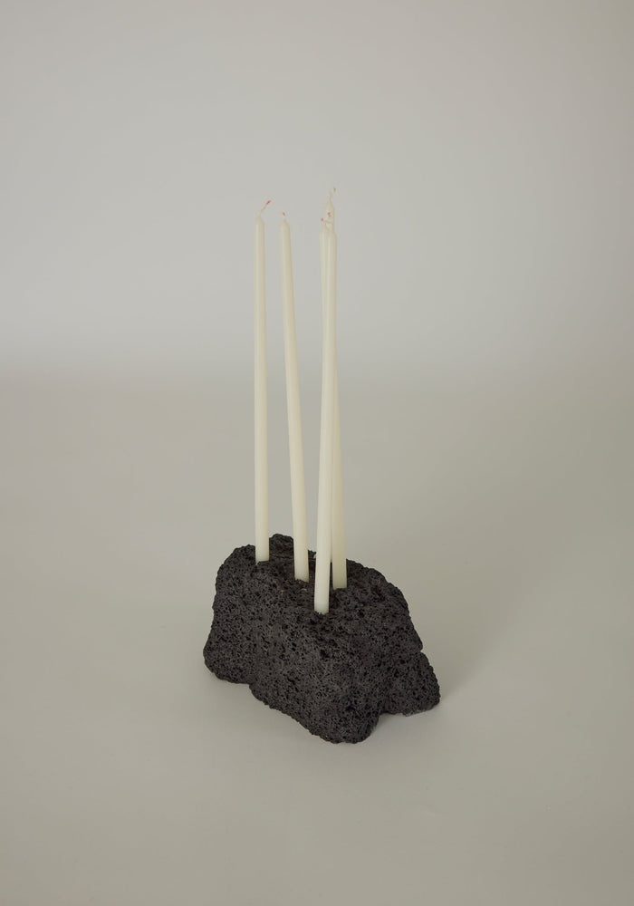 Sculptural Stone Candle Holder: A black, porous lava stone block with four white taper candles, handcrafted by Louis Crombag, showcasing minimalist Dutch design.