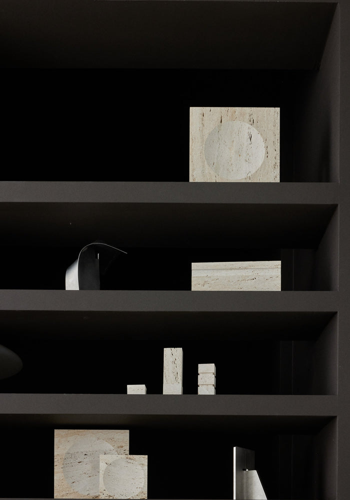 Medium tray | travertine displayed as a square stone block with a round indent, surrounded by minimalist geometric stone and metal objects on a matte-dark shelving unit.
