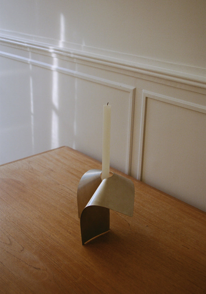 Stainless steel candle holder with a minimalist bent shape, featuring a brass-like finish, holding a white taper candle on a wooden tabletop. Handcrafted design by Paul Coenen.