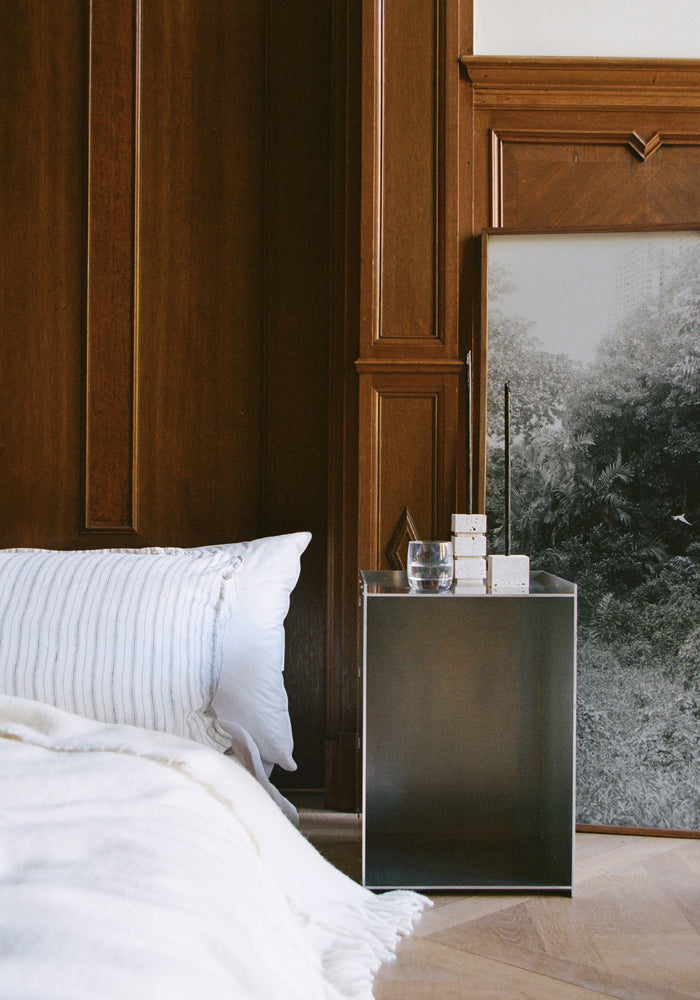 Two-way cube candleholder in travertine sits on a metal bedside table, showcasing its versatile design amidst a minimalist bedroom setting.