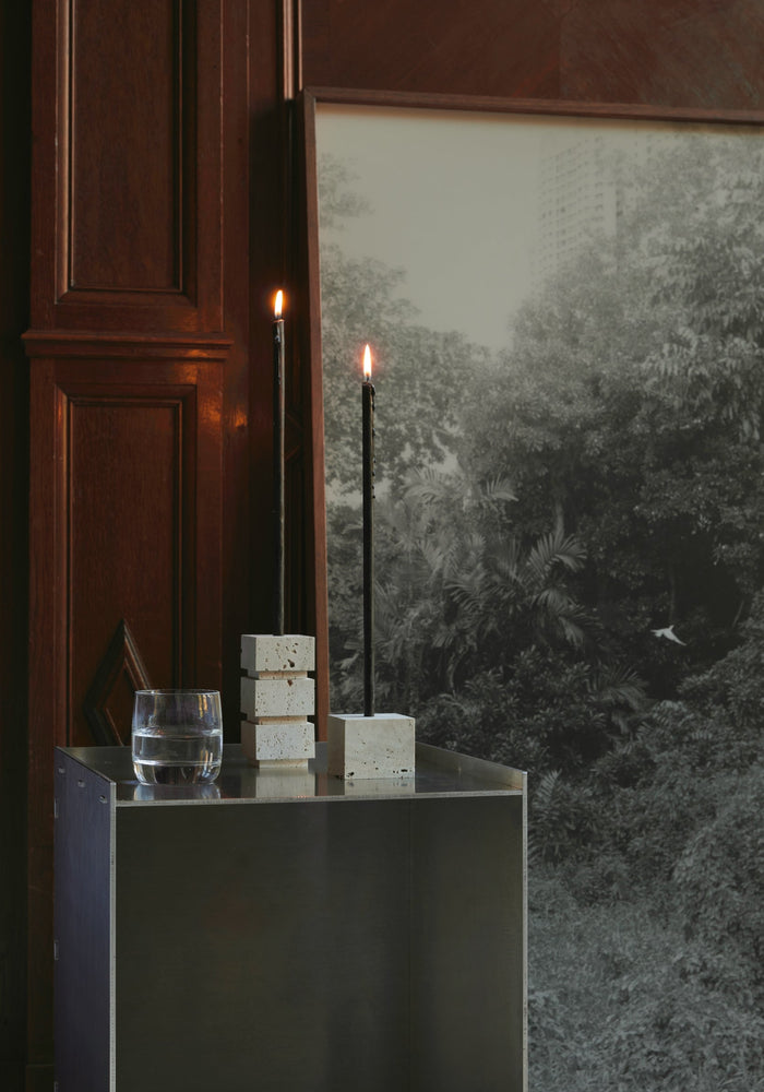 Sculptural candle holder small | travertine, featuring handcrafted stacked cubes and a single cube, holding two lit black taper candles on a metal pedestal.