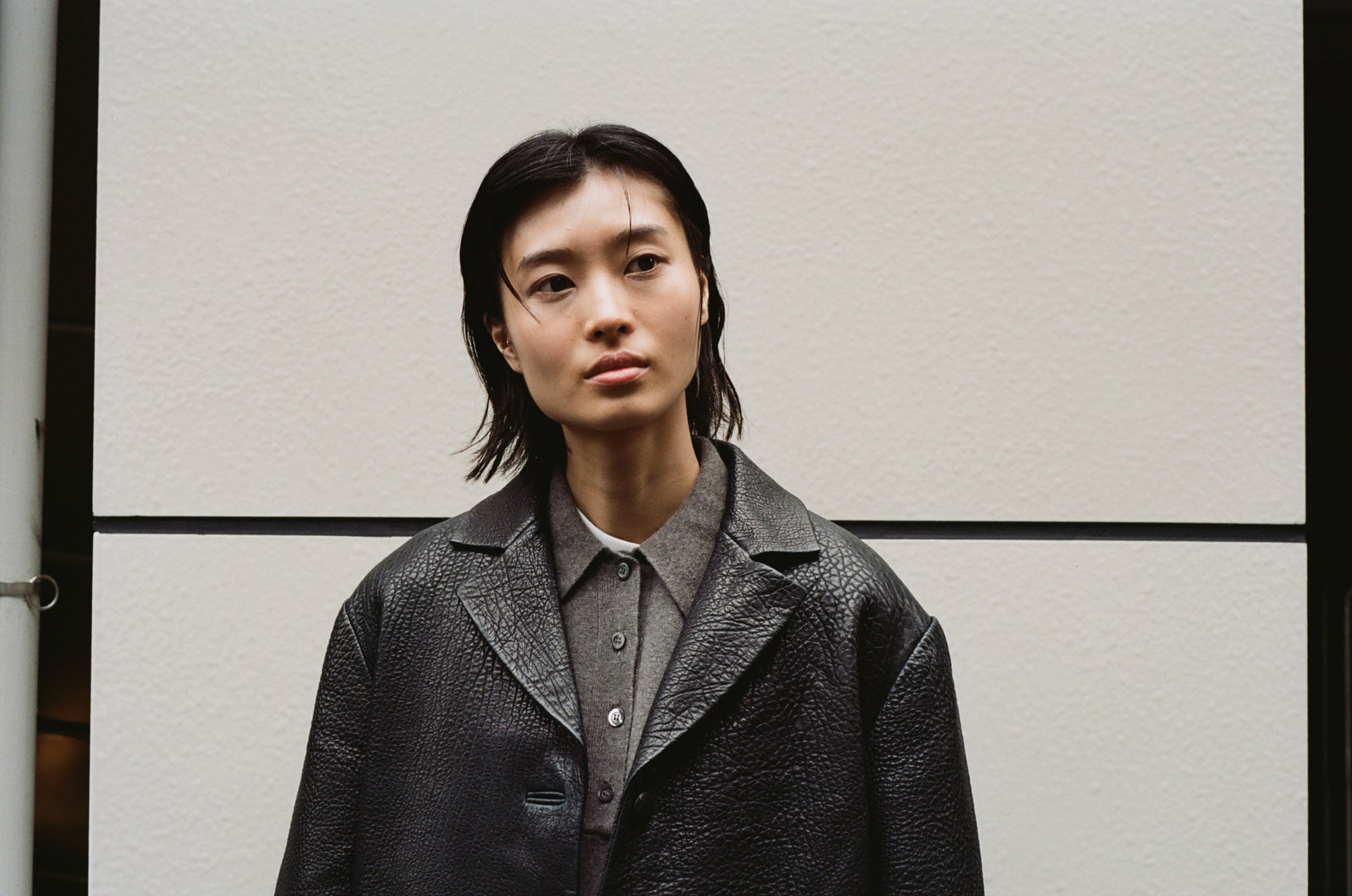 Woman with shoulder length black hair, wearing a textured leather blazer and a taupe polo against a white wall. 