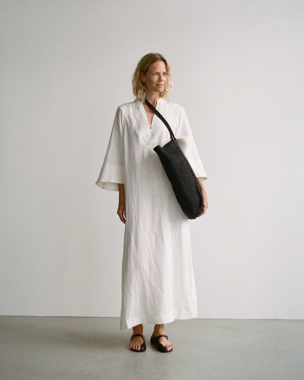 Woman in a white linnen kaftan dress holding a black straw bag against a plain background