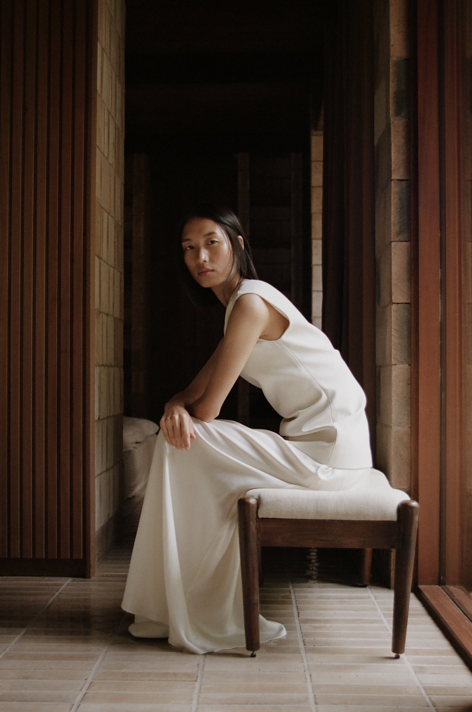 Woman wearing a long cream dress sitting on a bench in a interior with wooden walls and beige tiles