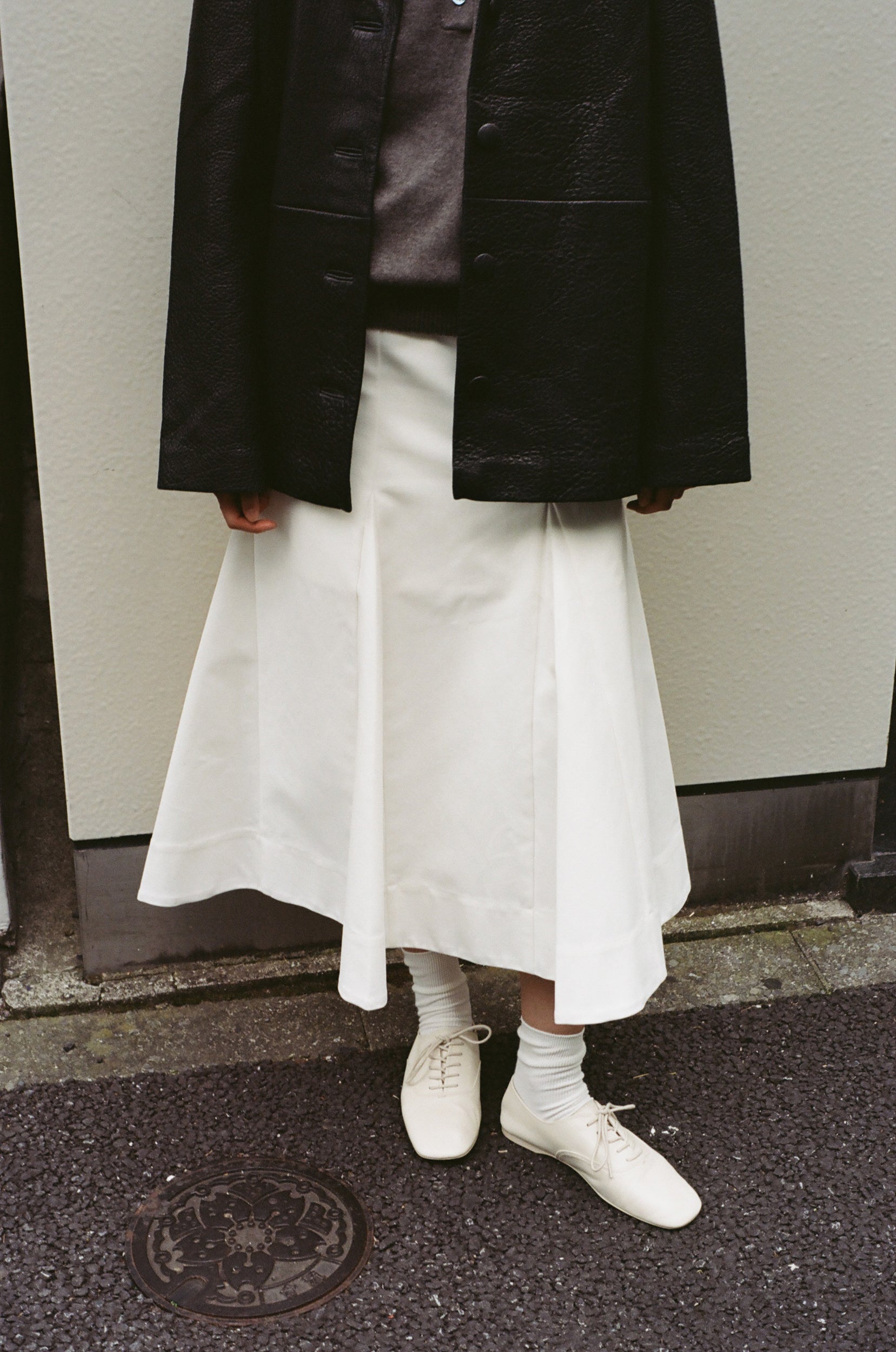 Woman wearing a leather blazer jacket and a cream sculptural skirt agains a clean wall in Tokyo. 