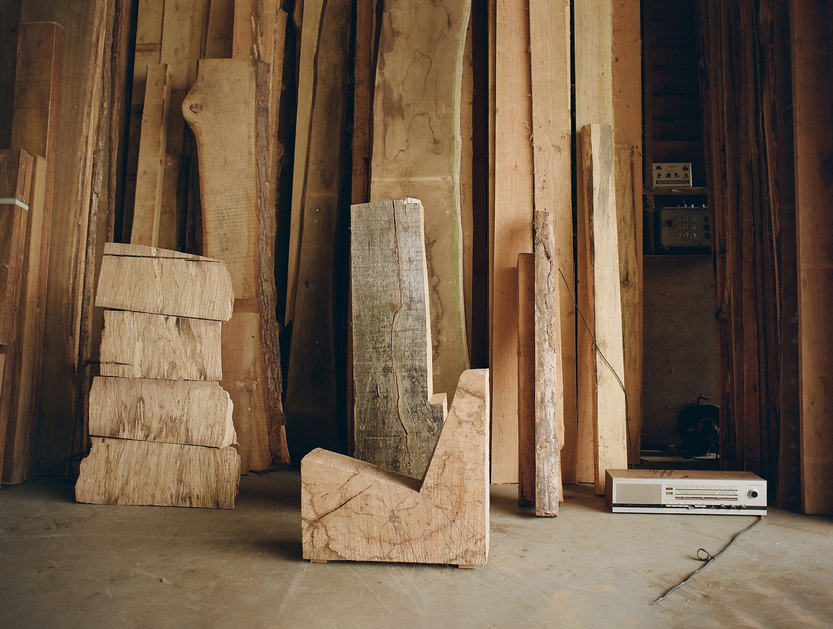 Stacks of wooden planks in a workshop setting with a sculptural shaped wooden chair in front and a 80's looking radio on the right