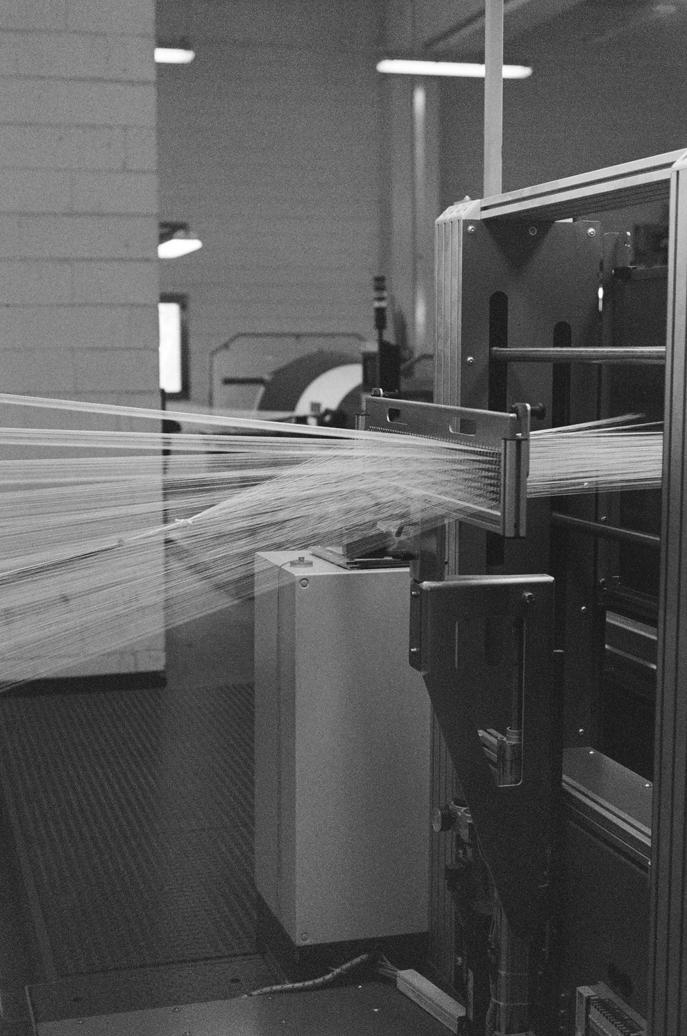 Black and white industrial setting with machinery and thin white threads being weaved together showing an intricate pattern