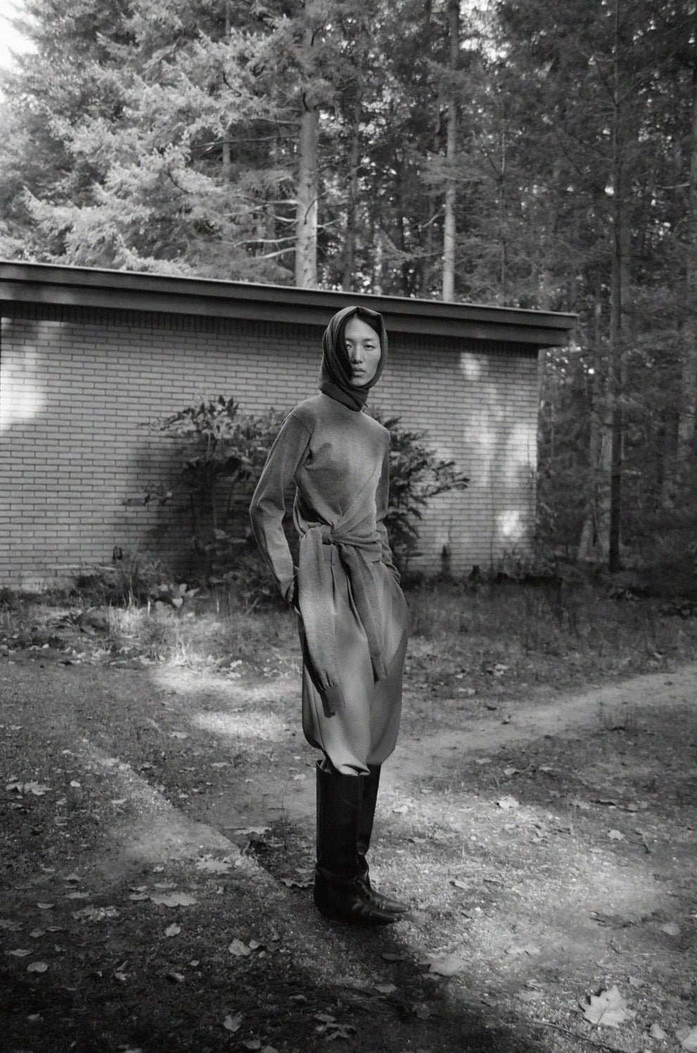 Woman wearing grey sweated and knitted scarf wrapped around her head in a forested area with a building in the background