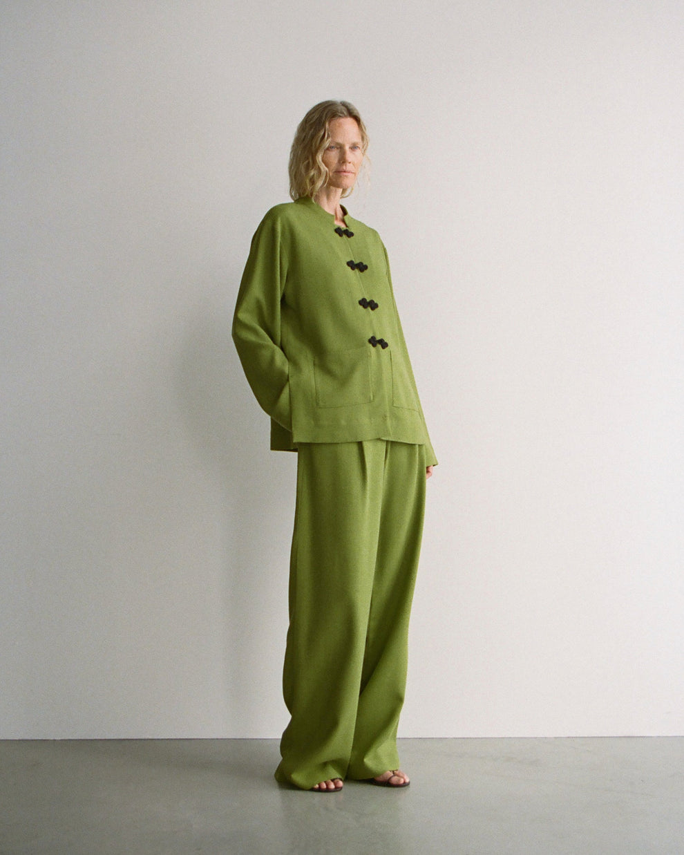 Woman wearing a green jacket with black mandarin closure and matching green wide leg trousers standing against a plain background
