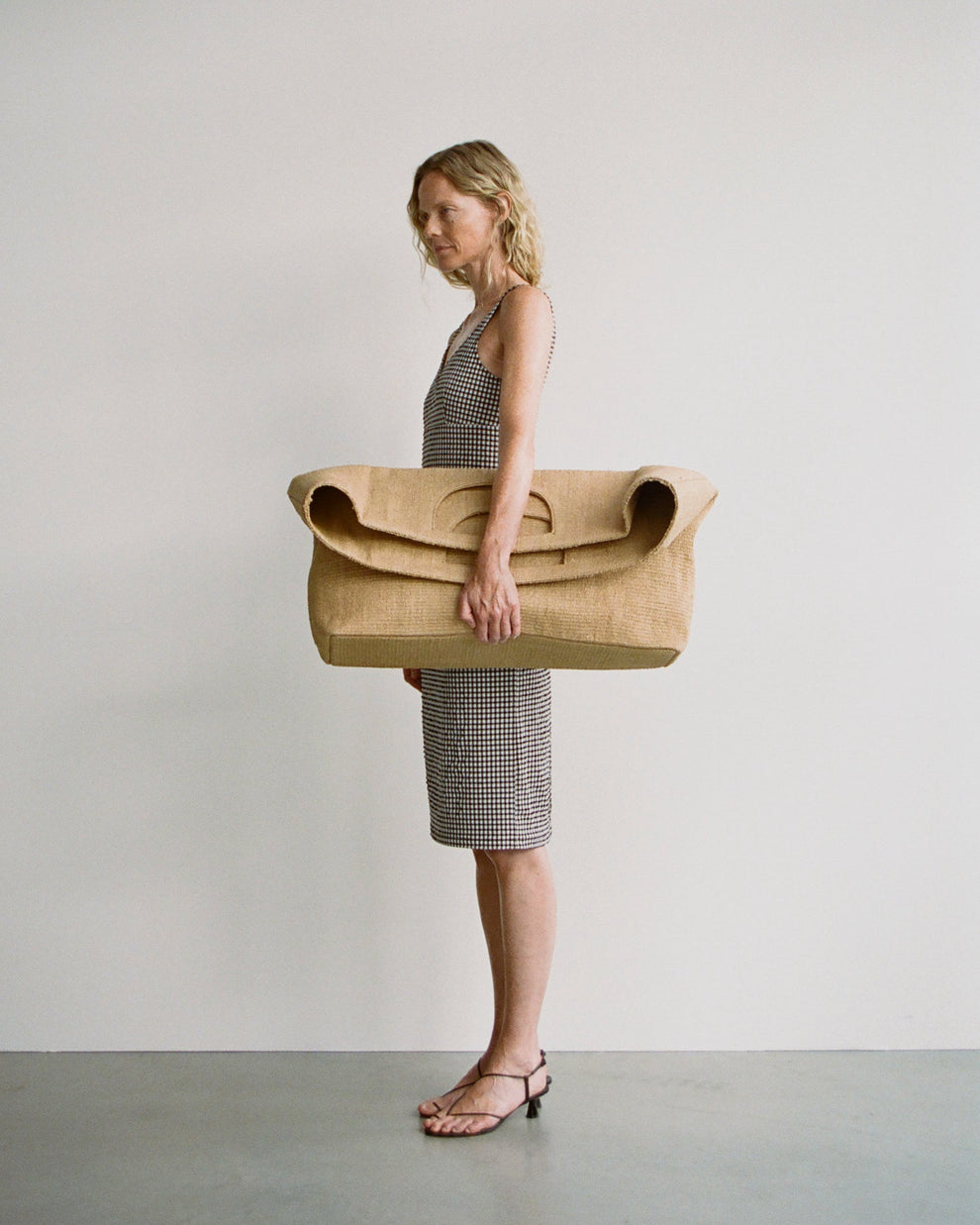 Woman wearing a fitted gingham midi length dress holding a large straw bag against a plain background