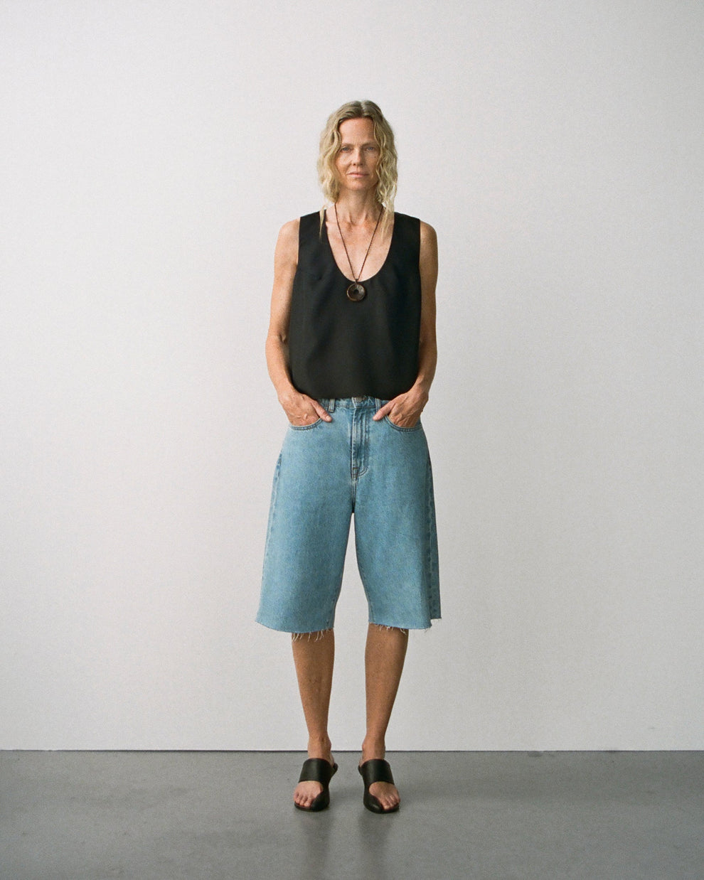 Woman wearing a black silk scoop neck sleeveless top and baggy denim shorts that go over the knee against a white plain background