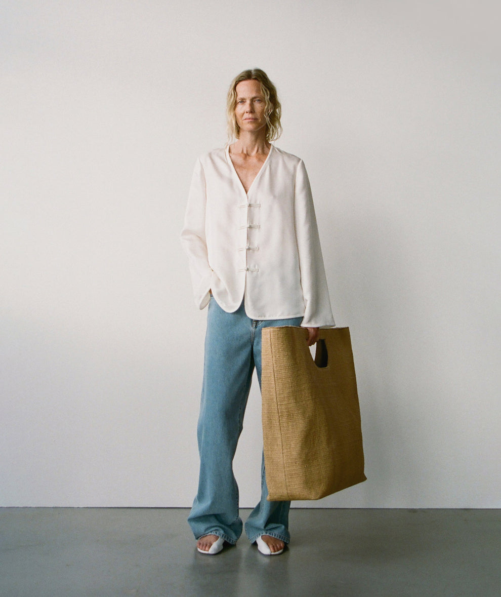 Woman wearing a cream silk top with v-neck and mandarin closure and baggy jeans holding a big straw bag against a plain background
