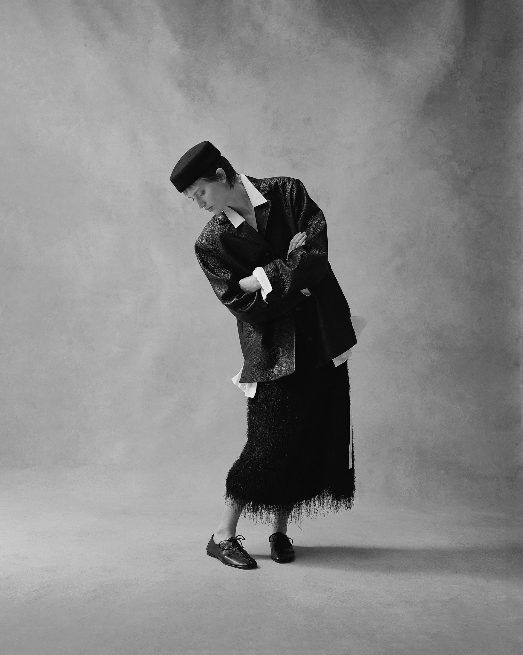 Black and white image of a woman wearing a black pillbox hat, black leather jacket, black fringe skirt, looking down against a neutral backdrop