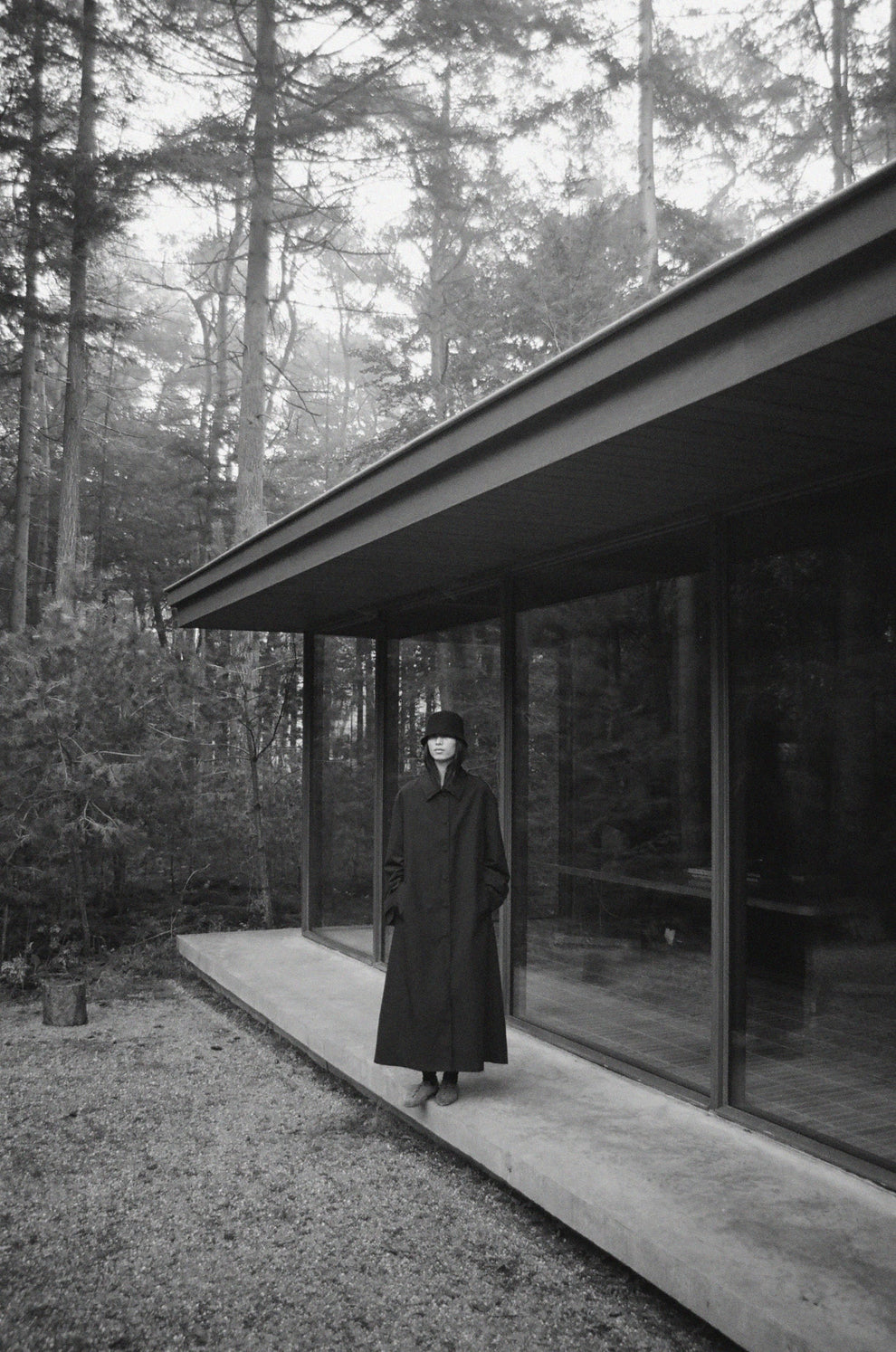 Woman in a long black trench coat and a black hat standing on a concrete deck of a modern bungalow surrounded by trees.