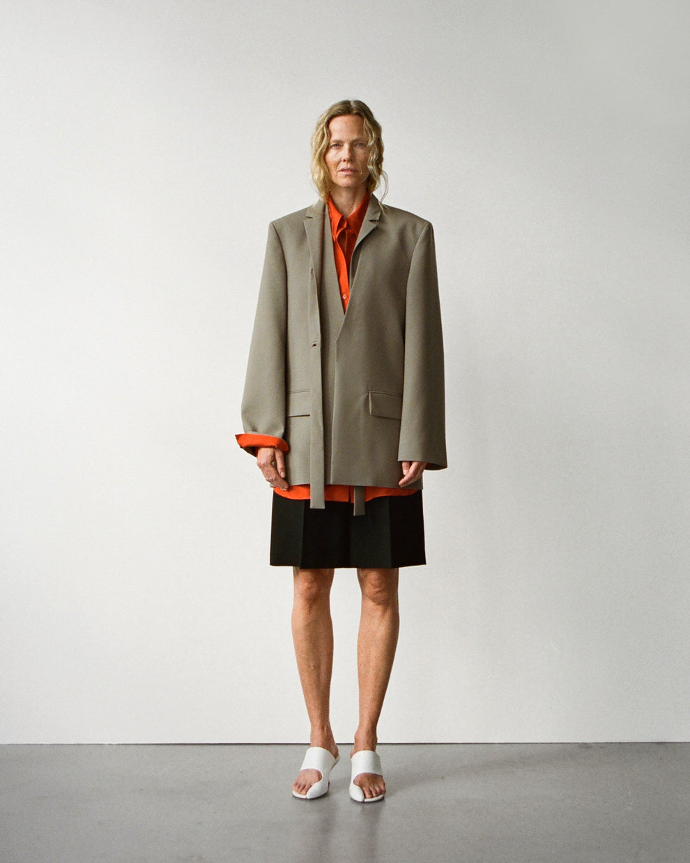 Woman wearing a taupe blazer over an orange shirt with black shorts on a plain background