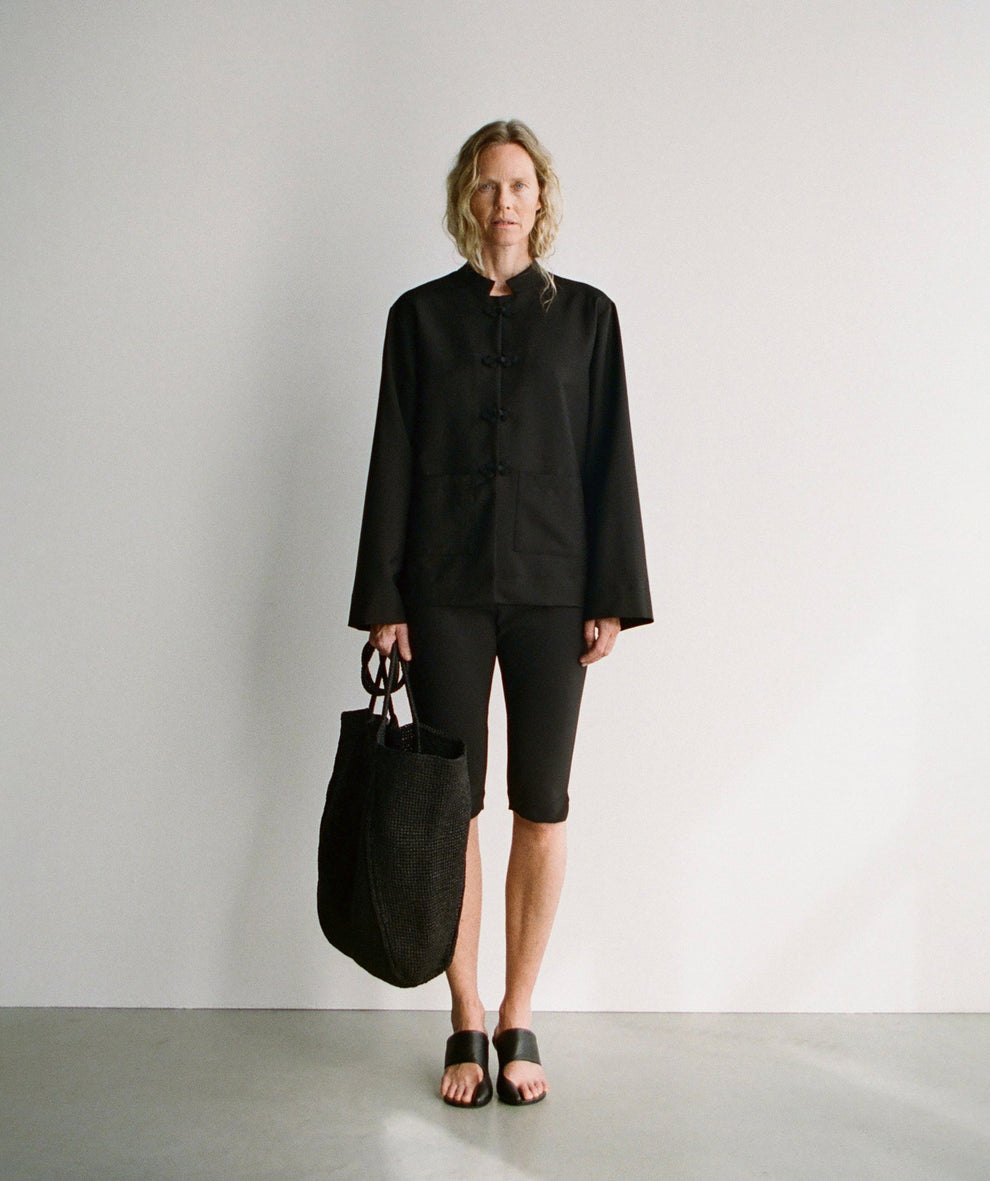 Woman wearing a black jacket with mandarin closure and black capri leggings holding a black straw bag against a plain background