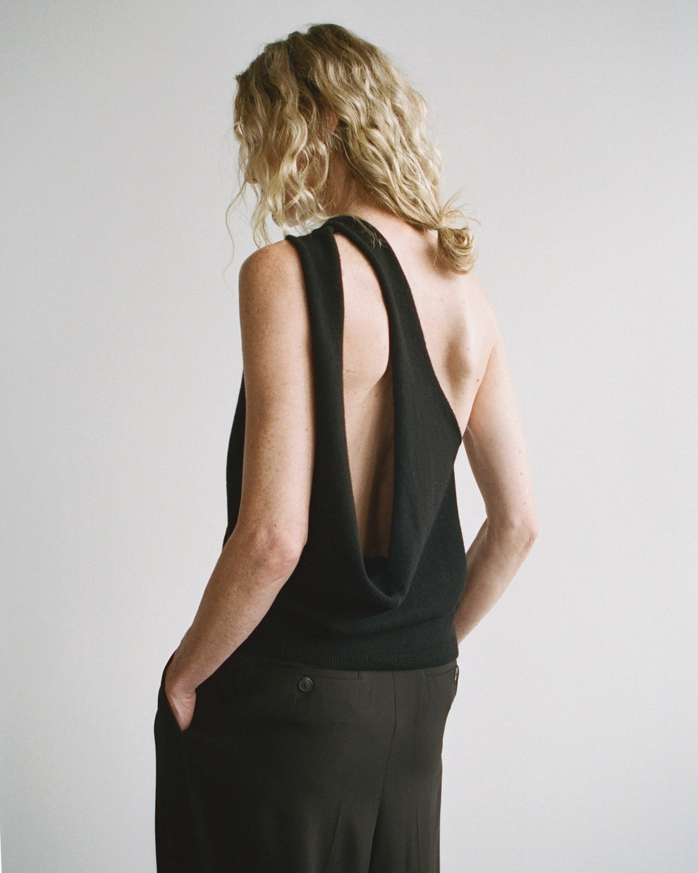 Woman wearing a black knitted one shoulder top with open back and her blonde curly hair in a low loose bun against a plain background