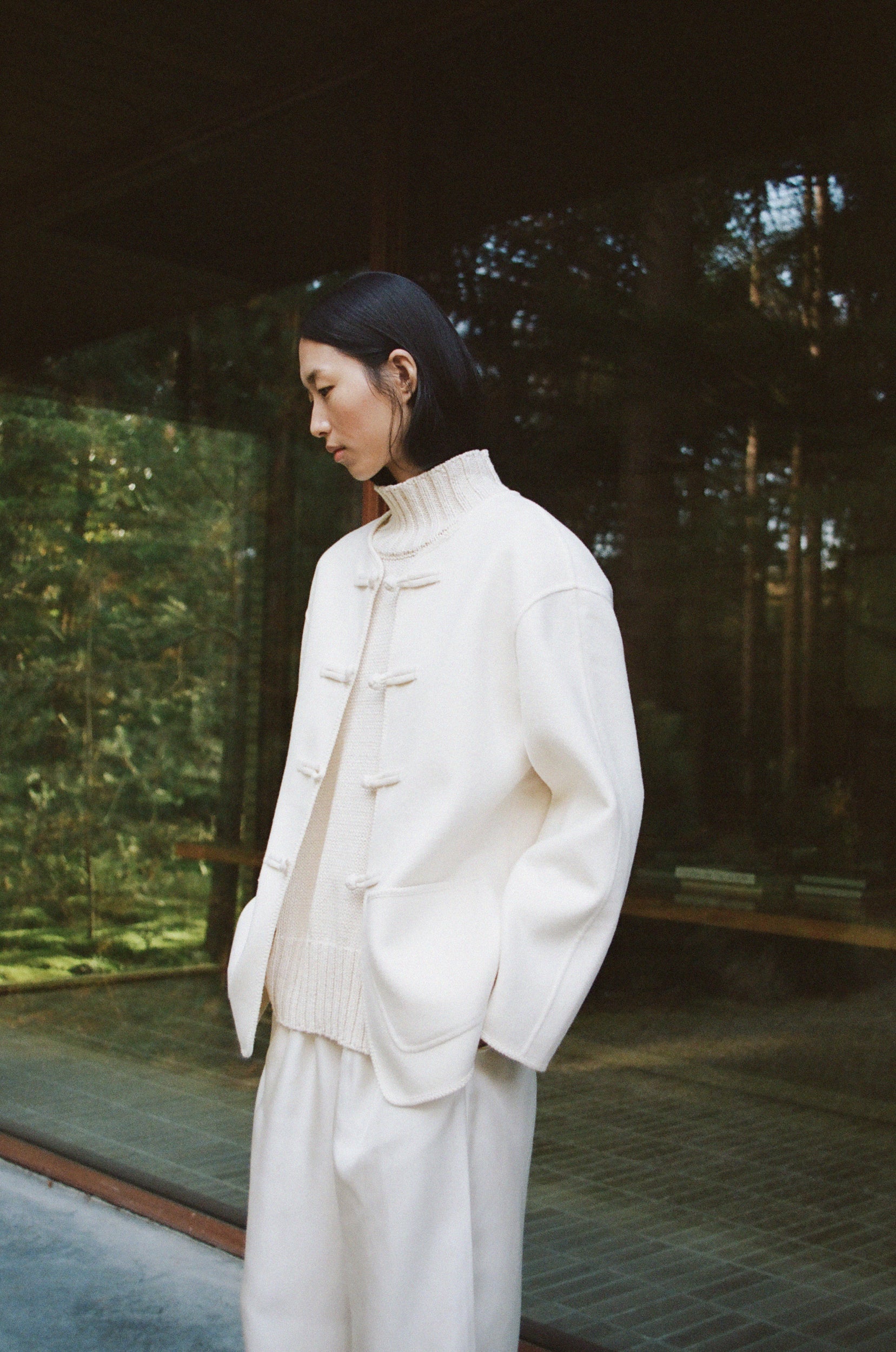 Woman wearing a cream turtleneck, cream double faced jacket and cream silk trousers standing in a serene outdoor setting with greenery reflected in a window.