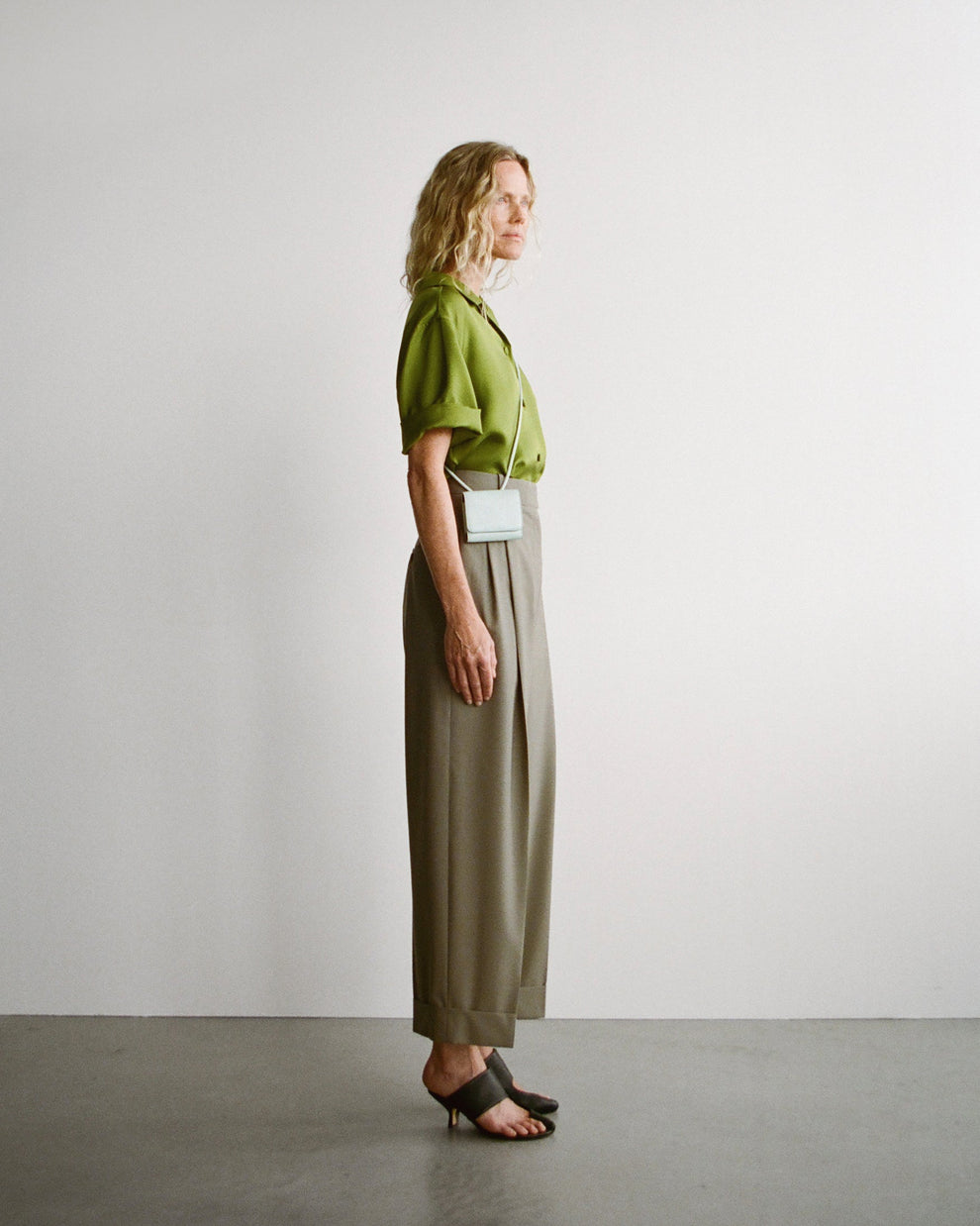 Woman wearing a green silk camp collar shirt and sage turn up pants standing against a plain white background