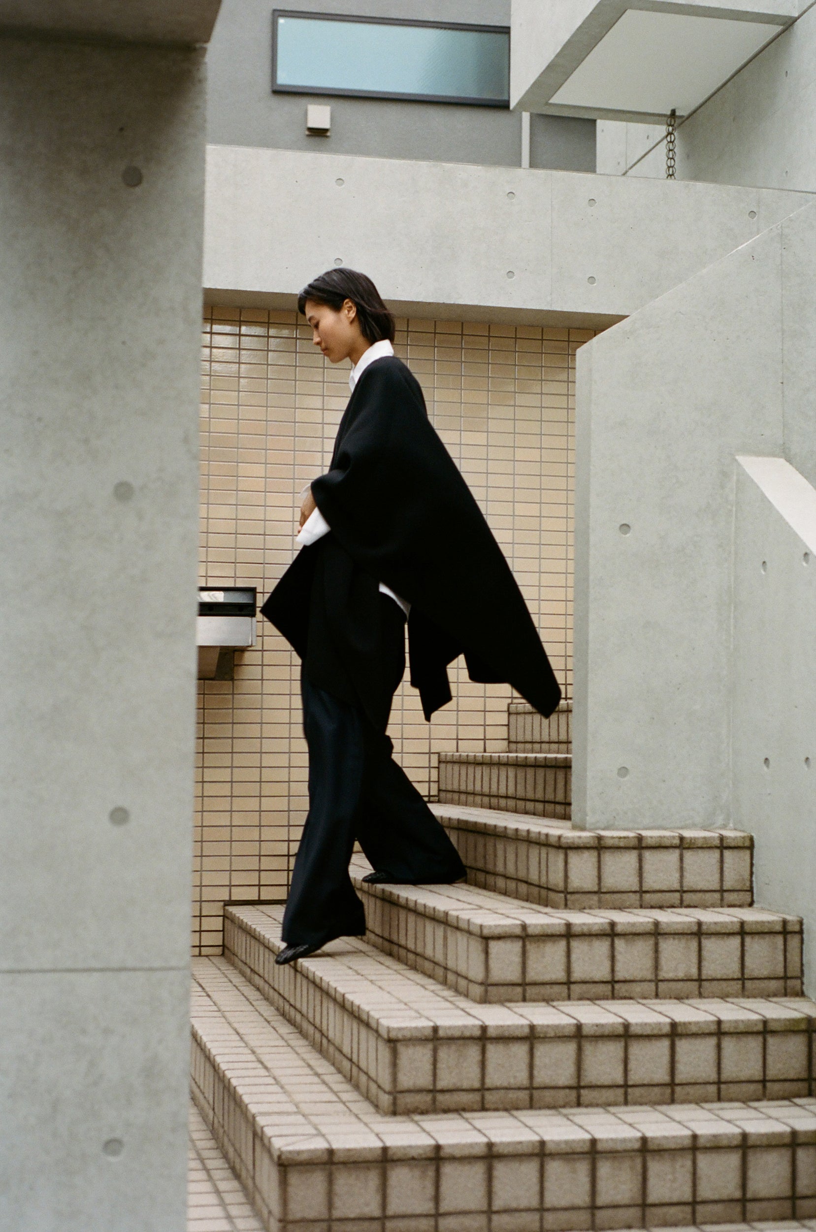 Woman wearing a black cape poncho, walking down tiled stairs in Tokyo. 