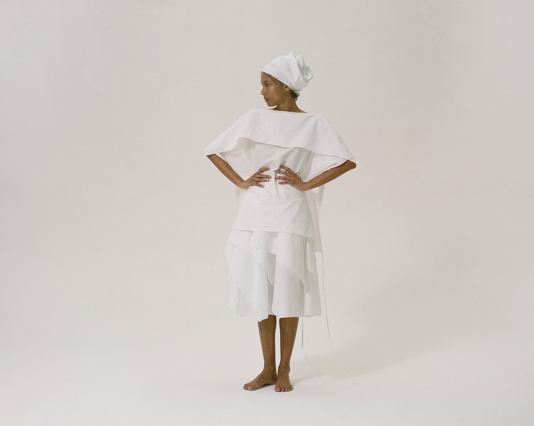 Woman wearing a white origami inspired cotton top and skirt with a white headscarf on a plain background