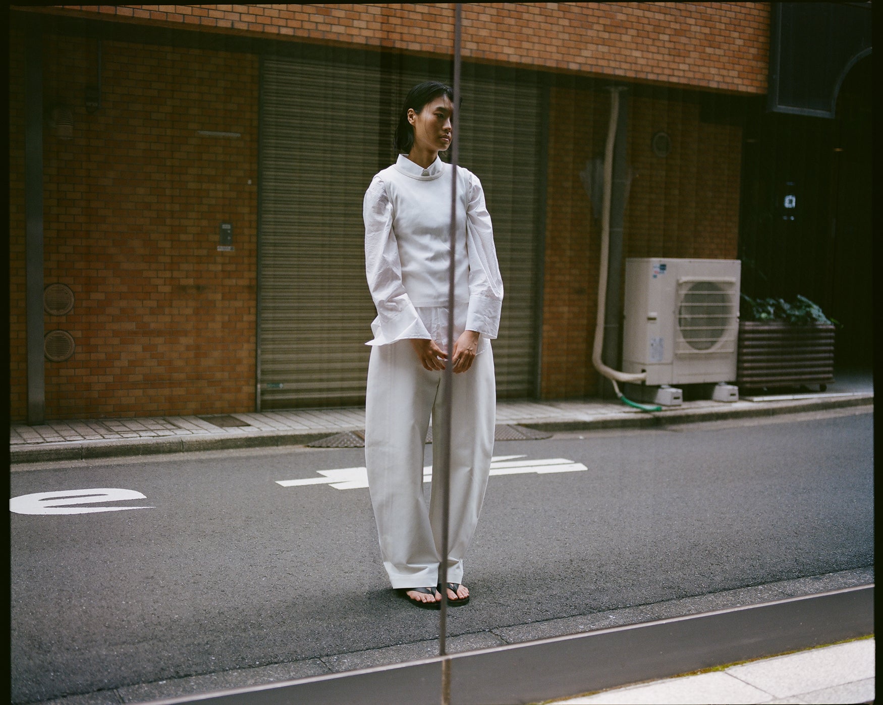 Woman standing in a street in Tokyo looking at herself in the reflection of a window, wearing a white outfit. 