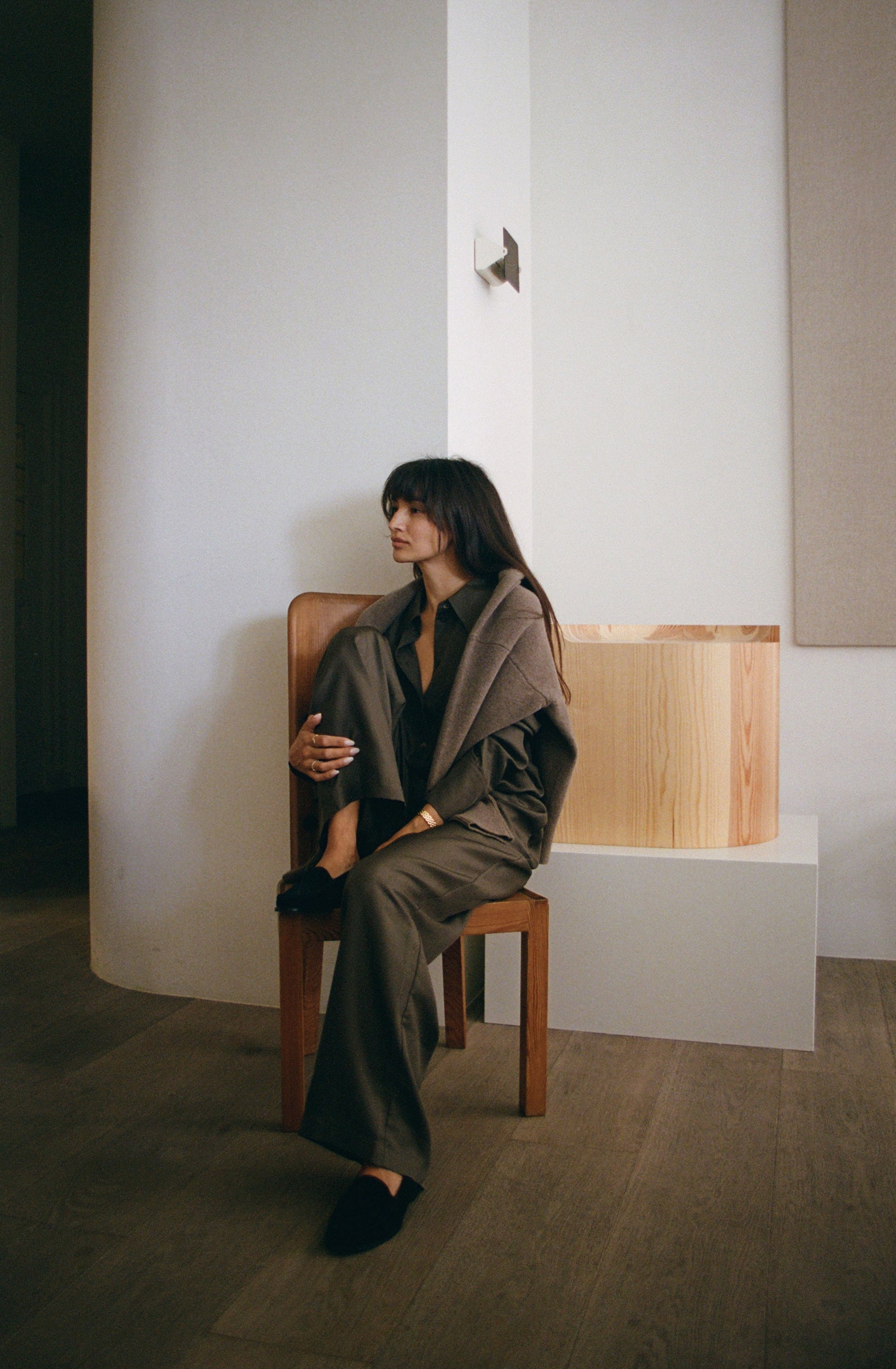 Woman sitting on a chair wearing a khaki green silk set in a minimalistic room.