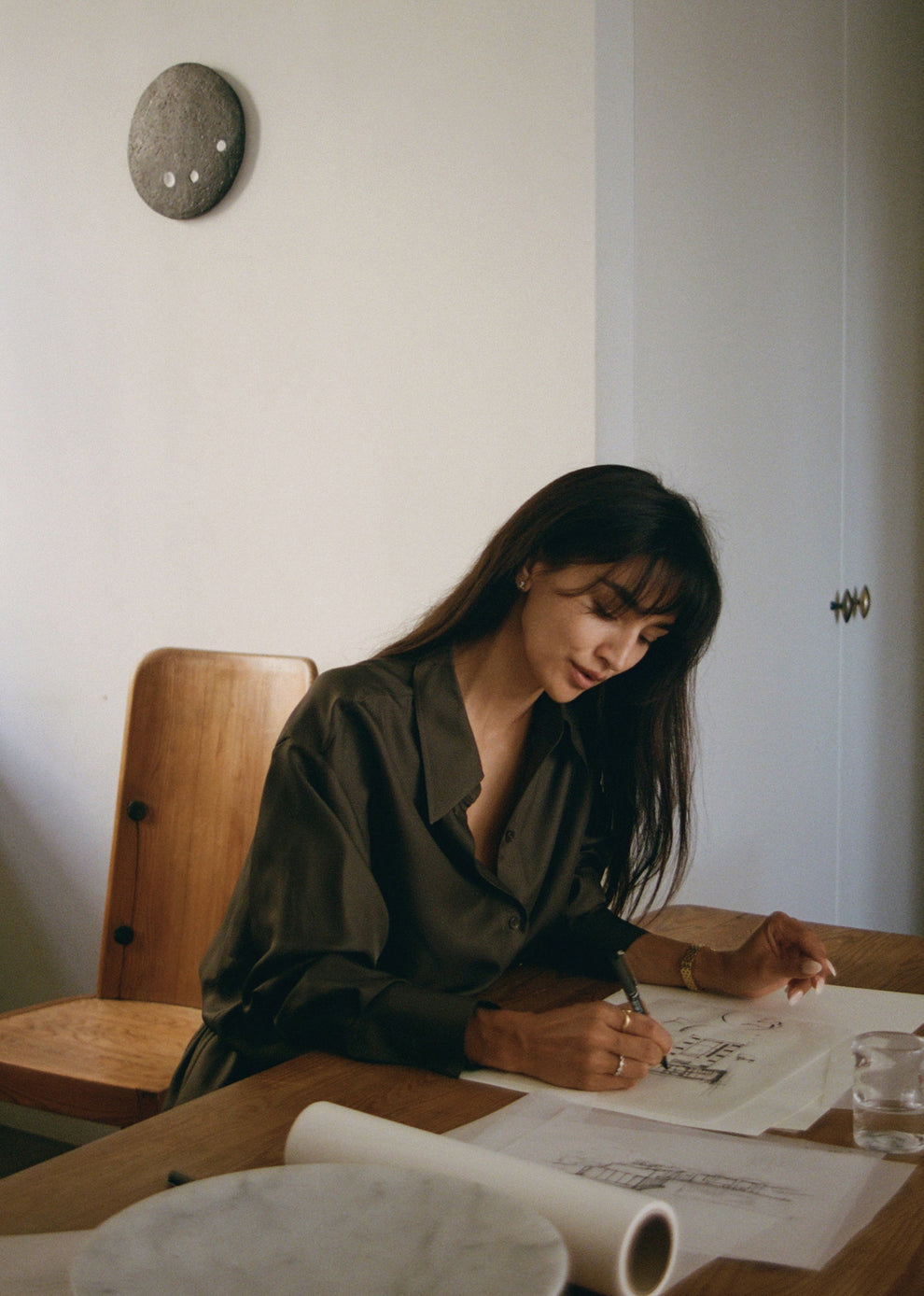 Woman sitting in a silk shirt at a desk drawing architectural drawings on paper with a pen.