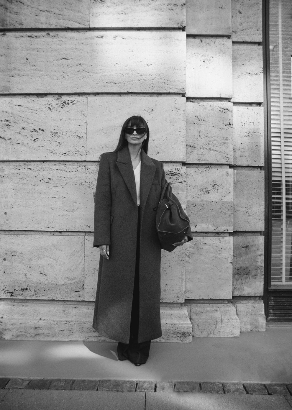 Woman wearing a long grey coat and sunglasses standing against a textured travertine wall holding a suede bag