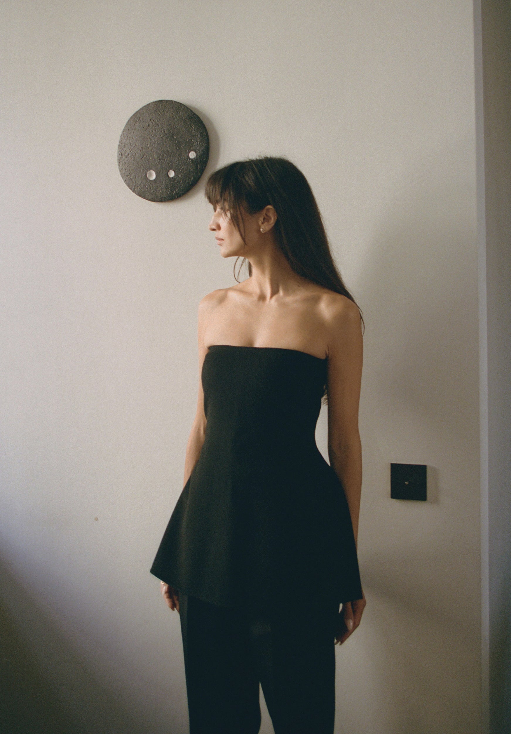 Woman wearing a black strapless top and black pants against a plain wall.