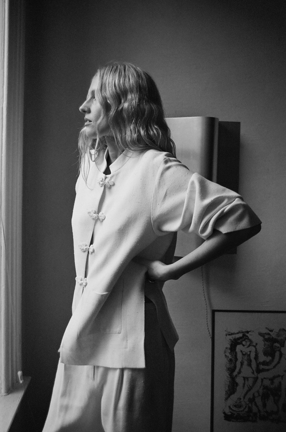 Woman wearing a white jacket with mandarin closure looking out the window standing against a white wall