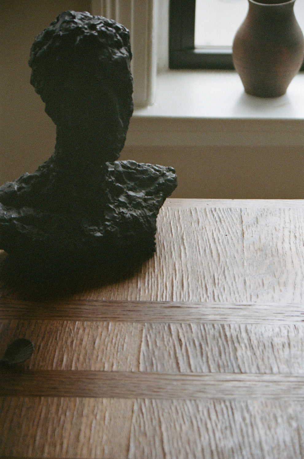 Dark stone sculpture of a woman's head on a wooden table with a vase in the background.