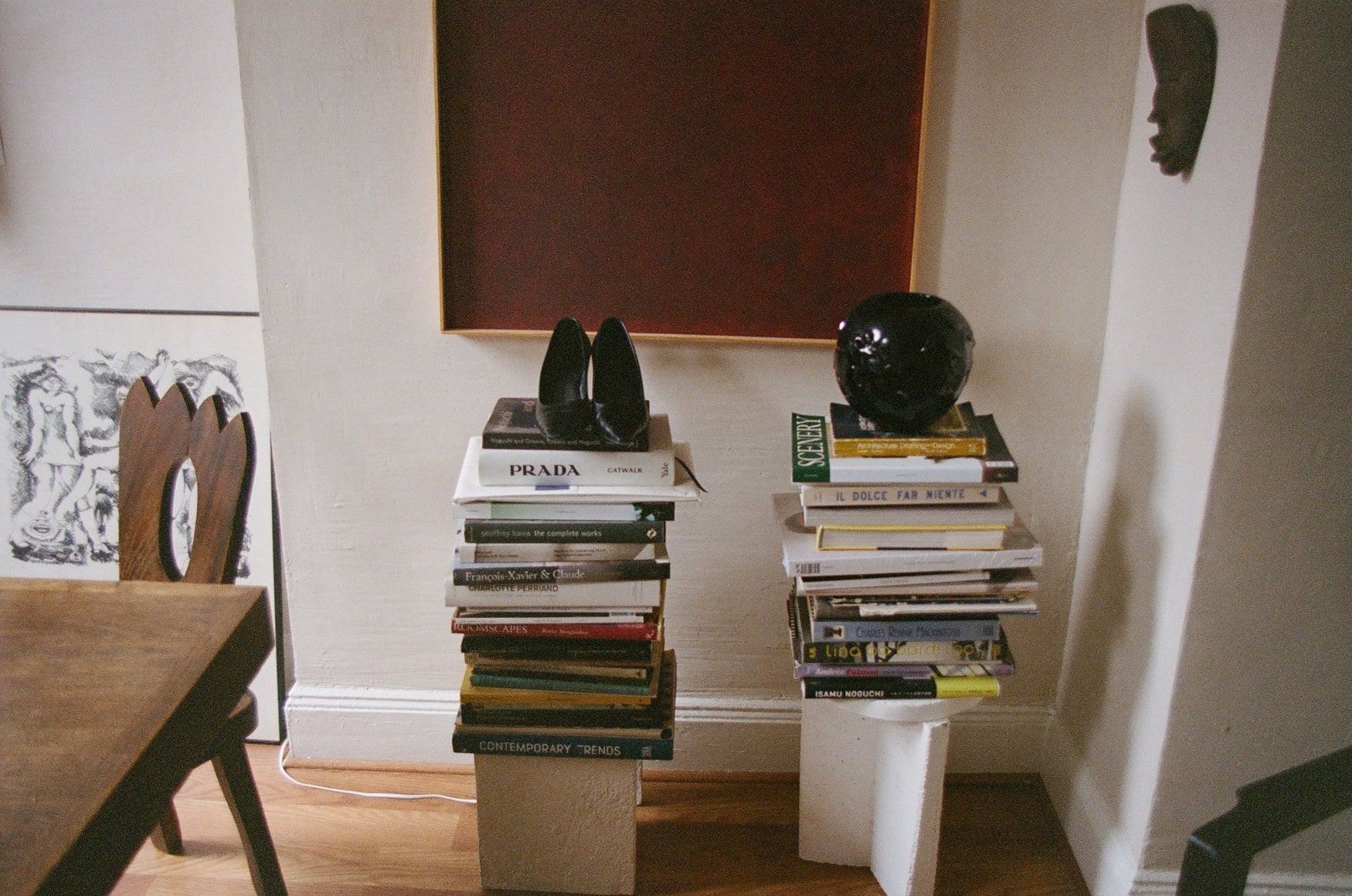 Stacks of books on small tables against a wall with a wooden floor with shoes and a vase placed on top