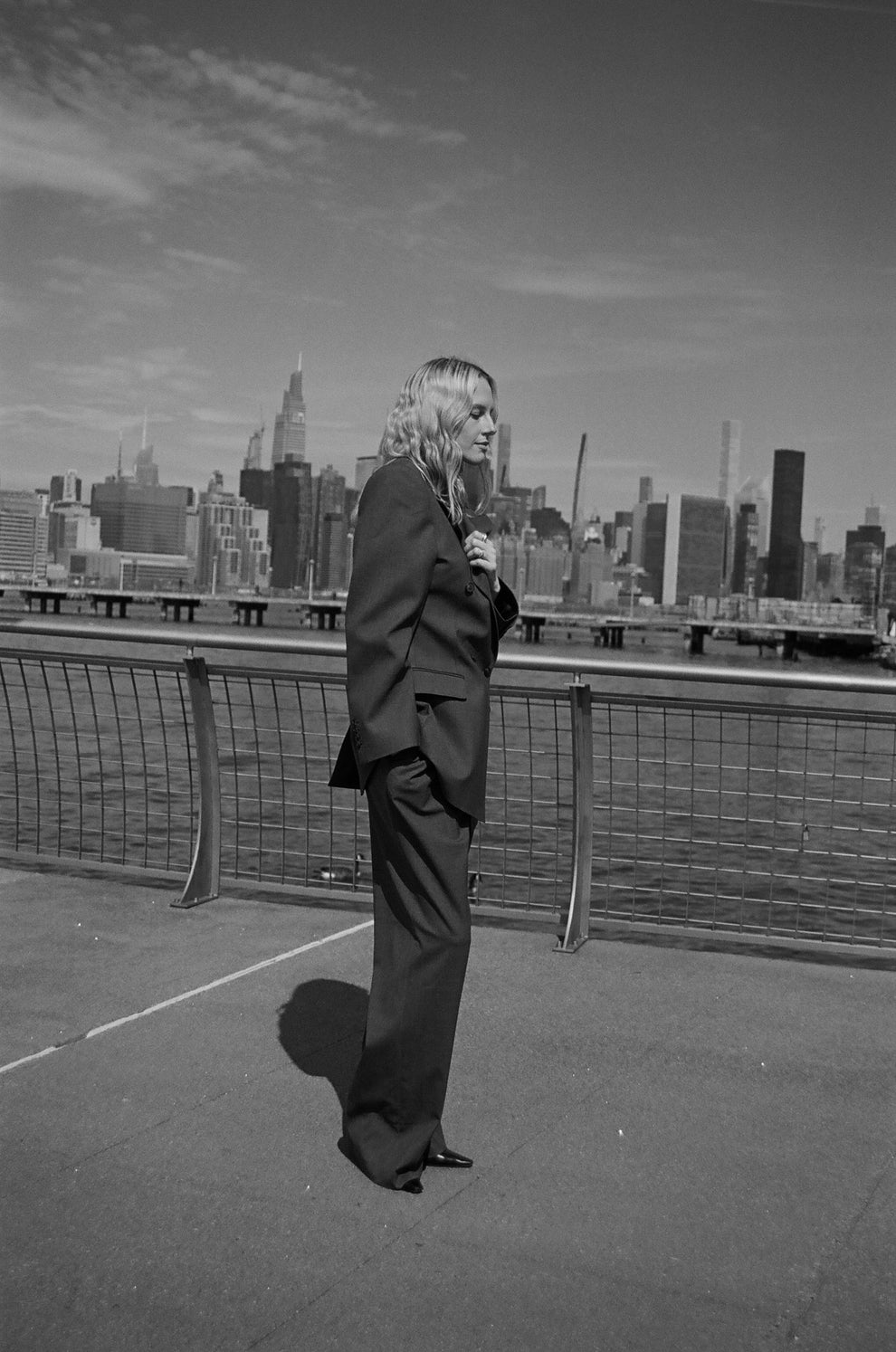 Woman in a grey suit standing next to the Hudson river with the New York city skyline in the background