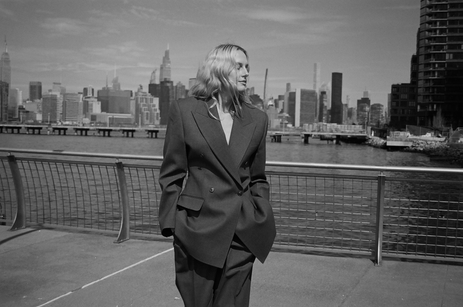 Woman in a grey tailored suit standing next to the Hudson river in with the New York city skyline in the background