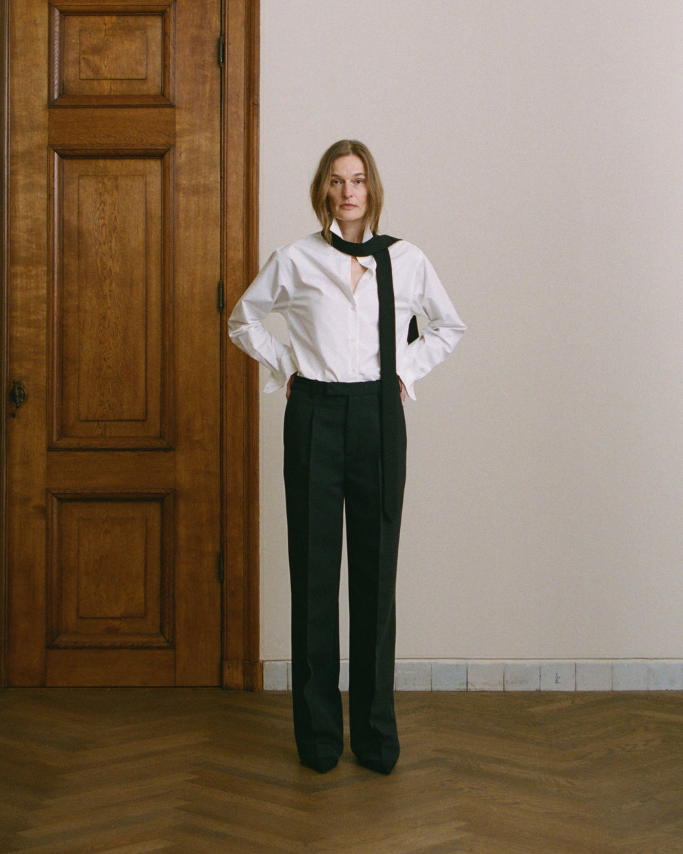 Woman wearing a white blouse with a black tie and black pants standing in a room with wooden floor and wooden door.