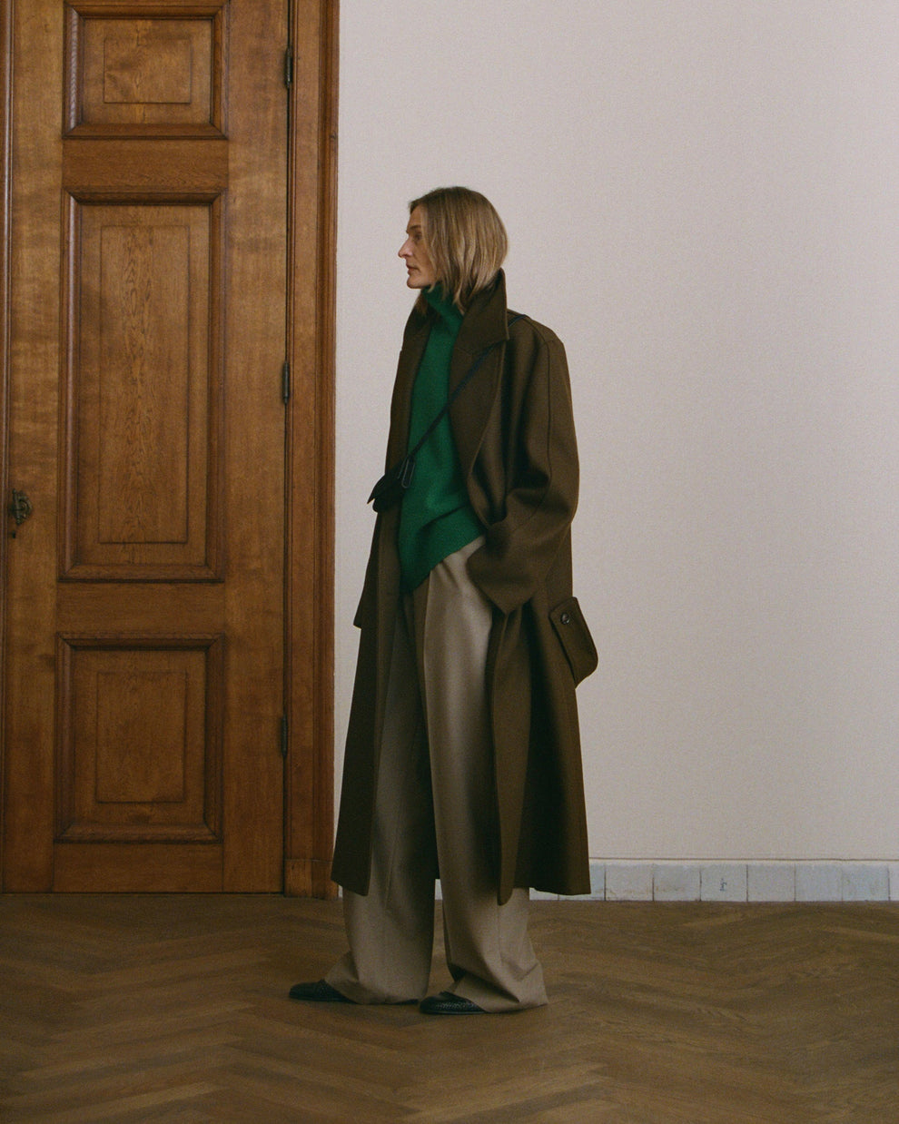 Person wearing a long dark brown coat and green sweater standing in a room with wooden floor and white walls.