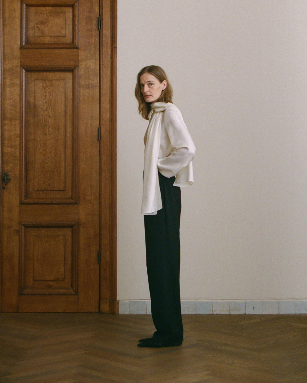 Woman wearing a cream silk top with scarf detail and black trousers standing in a room with wooden door, wooden floors and a white wall