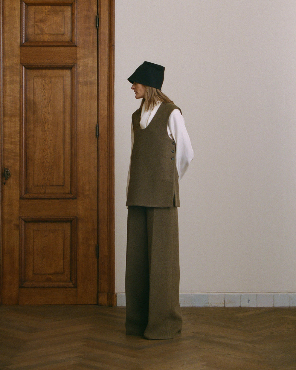 Woman wearing a green vest layered over a cream silk long sleeve top and matching green pants standing in a room with wooden floor and white wall.