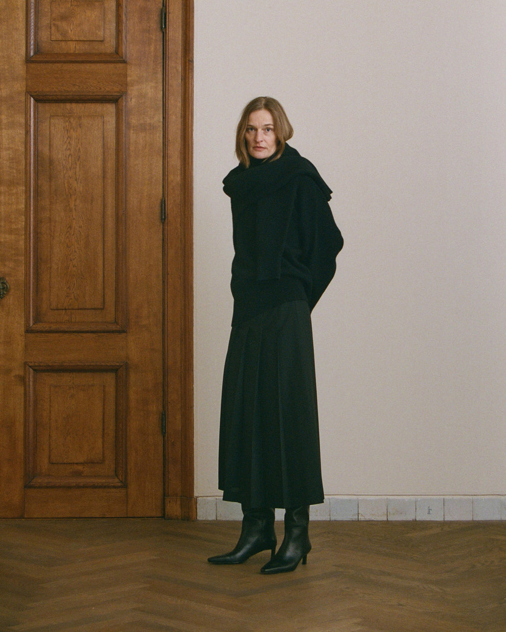 Woman in a black turtleneck sweater and black pleated skirt standing in a room with wooden doors and floor and a plain wall.