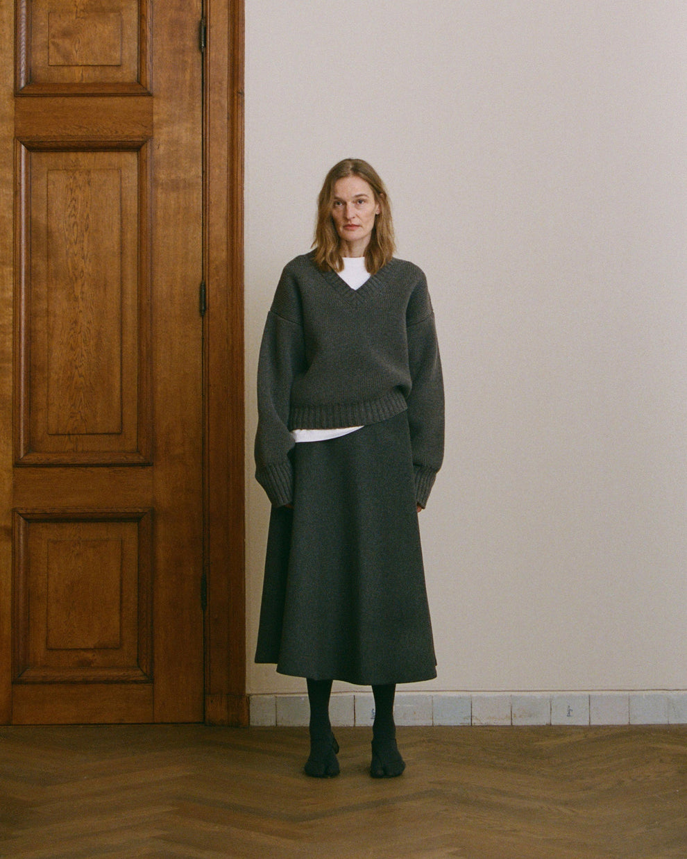 Woman wearing a dark grey thick v-neck sweater and dark grey a-line wool skirt standing in a room with wooden floor and paneling.