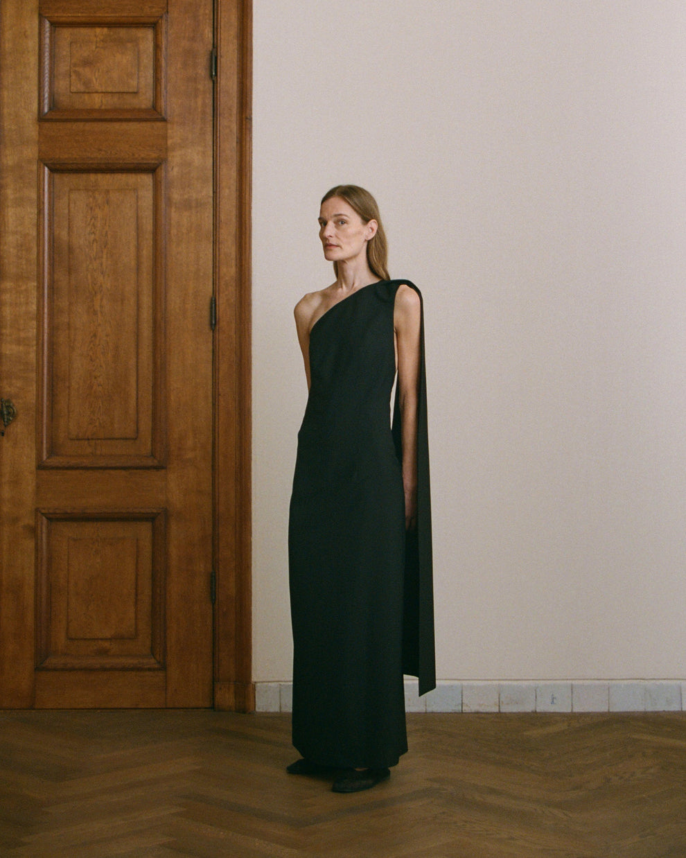 Woman wearing a black one-shoulder dress with scarf detail hanging down standing in a room with wooden flooring and a wooden door.