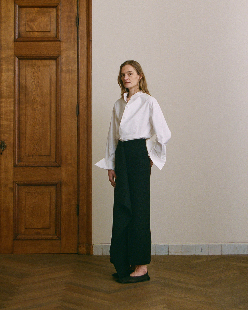 Woman wearing a white blouse and black long skirt standing in a room with wooden floor and door.